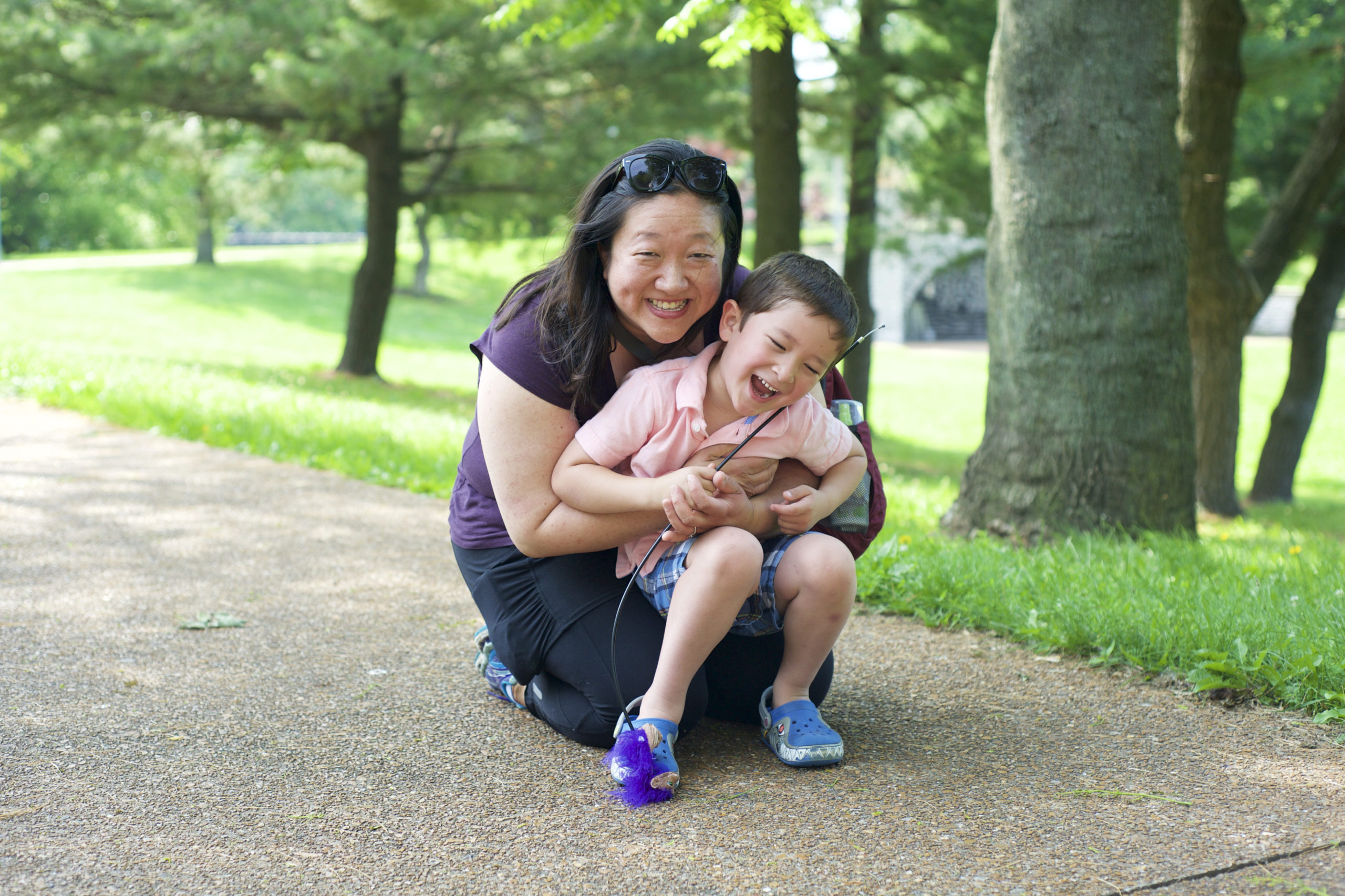 A photo of Lisa Harper-Chang and son crouching on the ground in a tree-lined park with green grass. Lisa is an East Asian woman with sunglasses on her head and shoulder-length black hair and is wearing a purple short sleeved top. Her child is smiling and laughing in her arms and sitting in her lap as she is kneeling. The child has light brown short hair, a button up pink shirt, blue shirts, and blue shoes.