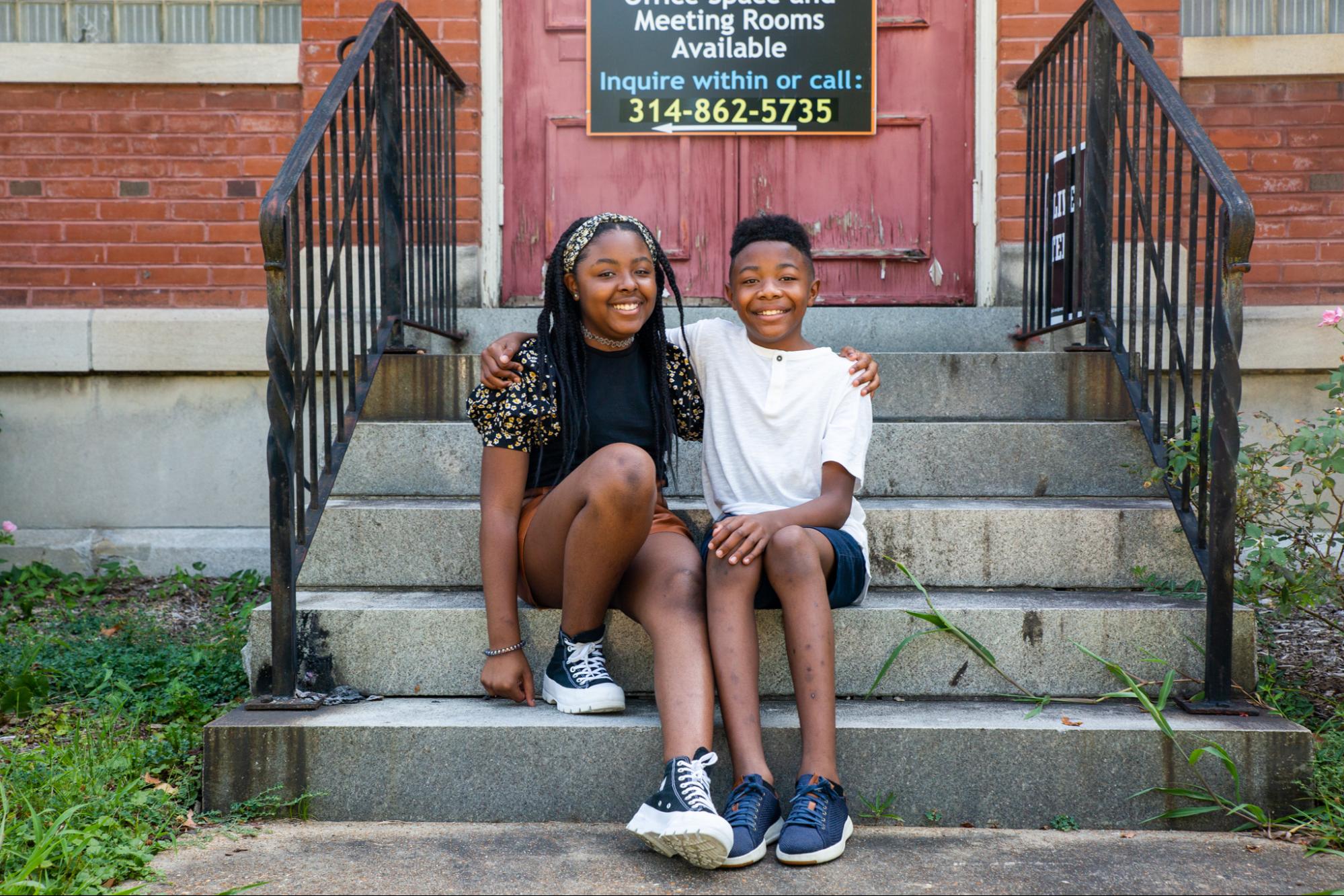 Sister and brother Grace and Yosiyah, Black siblings, Yosiyah in white t-shirt and a grin and arm around his sister, sitting on the front steps of the building for the American Friends Service Committee