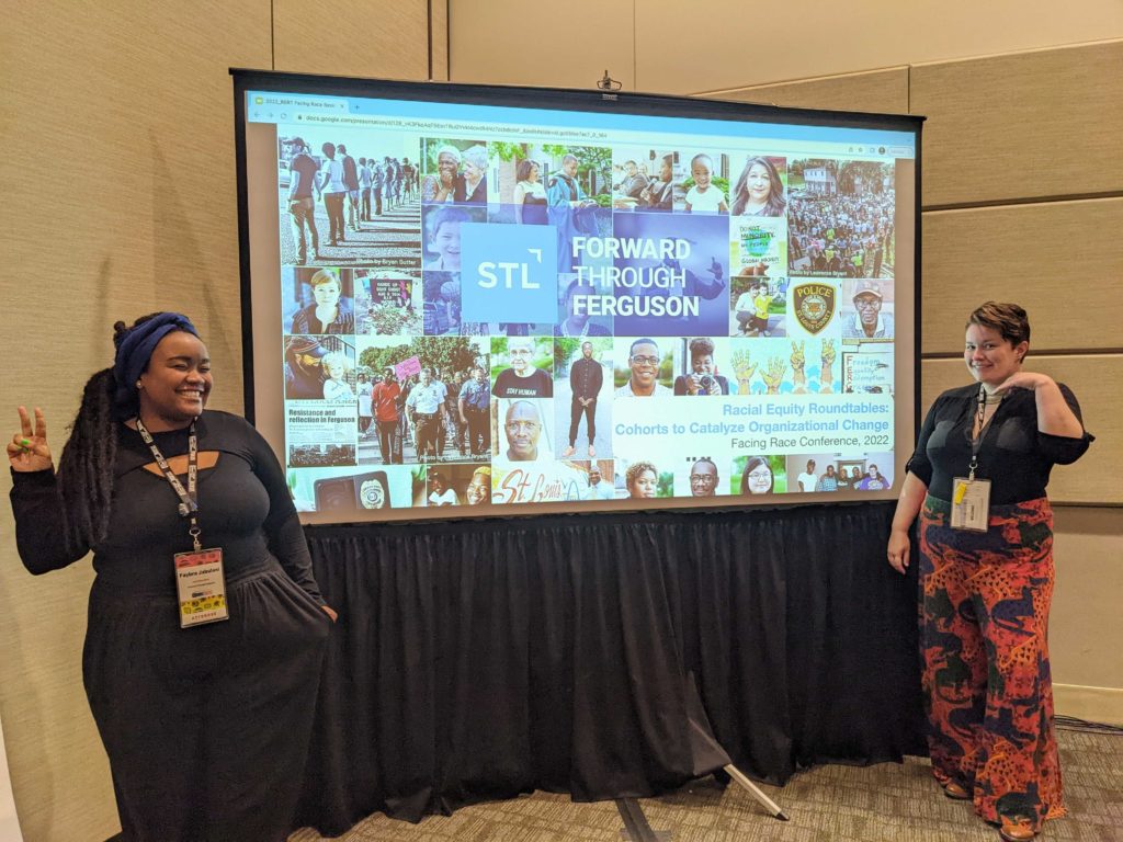Faybra (a Black woman putting up a peace sign) with lots is smiling and Sarah (a white nonbinary femme) is putting their hand under their face and smiling. They are standing next to their Racial Equity Capacity Building presentation at the Facing Race 2022 conference workshop.
