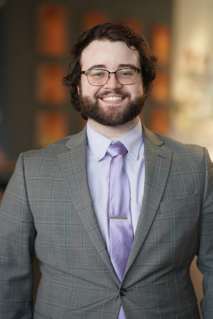 Image of Stephen Jung, a white man with brown curly hair and a brown beard. He is wearing wire frame glasses and is smiling in a grey suit and purple button up with purple silk tie. 