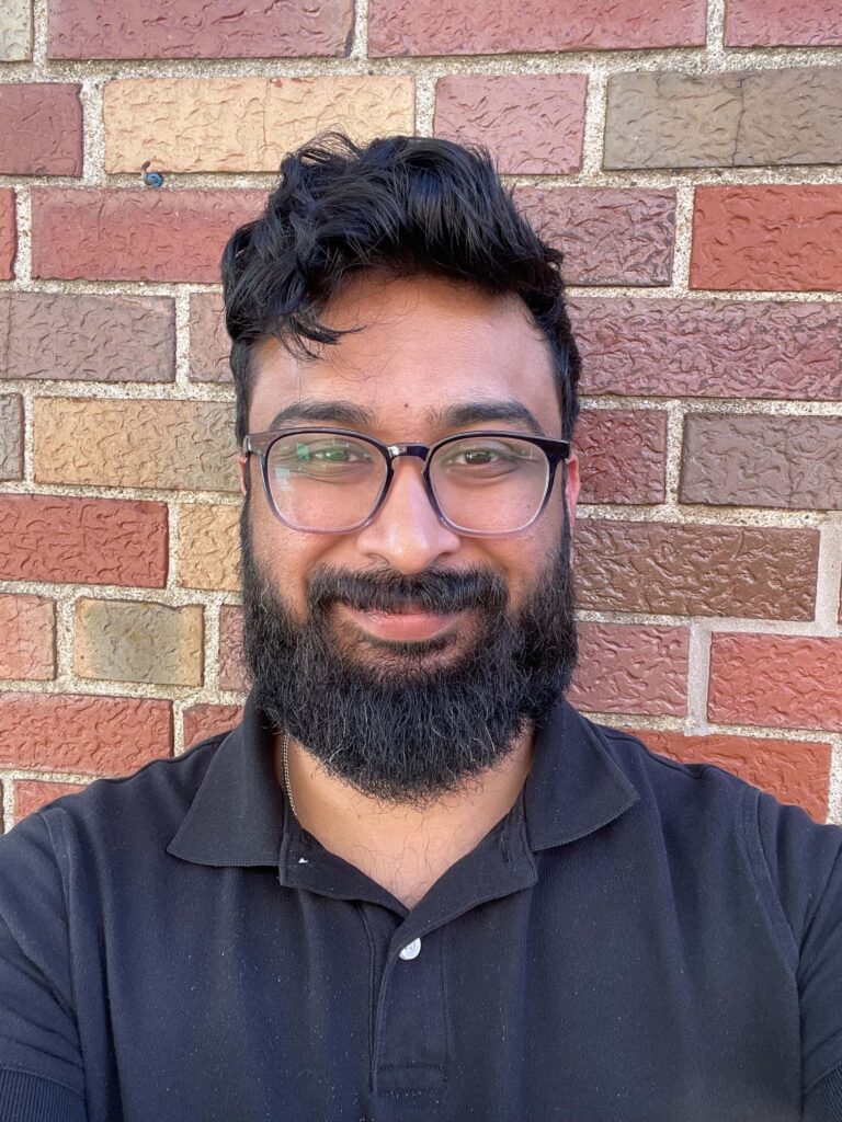 Madhav Narayan, Forward Through Ferguson's Senior Director of Policy & Data, is pictured from the shoulders up against a brick background. He is wearing a navy shirt and has a thick black beard and a short haircut with thick curls at the top. He is wearing glasses and has a closed mouth smile. 