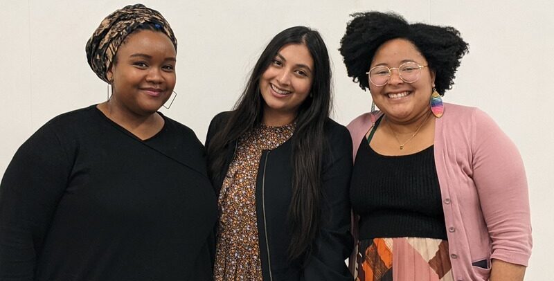 Photo of Faybra, Lead Racial Equity Capacity Catalyst on left, Sara, Lead Catalyst, Resource Development in the middle, and on right Nichole, Director of Community Partnerships. Faybra is a Black woman in a black shirt and with a scarf around her head who is smiling with her mouth closed. Sara has long black hair, a black cardigan, and flowery top and is tilting her head to the right smiling with teeth. Nichole has natural hair parted on the side, with round glasses, colorful earrings, pink cardigan, black top inside, and colorful skirt.