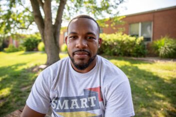 Josh Jones is a Black man sitting under a shady tree with bright green leaves dappled with sunlight in the background. He has neat close cropped haircut and a close cut beard. He is wearing a t-shirt that says SMART in all caps letters at the top.