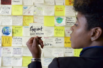 De Nichols a Black woman whose back is to the camera, with her face turned to the left and her hand holding a sticky note say that the true expert is he/she who is affected most by the issues. There is a wall of sticky notes in front of De. De is wearing a silver watch, a navy blazer, and has natural hair with more length at the top rising up from her head.