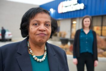 Photo of Jacqueline Hutchinson, an older Black woman with a bob. She has a closed mouth smile, is wearing a navy blue blazer and turquoise top along with a multi-colored beaded necklace. In the background is her colleague, Elisabeth Risch, who is a white woman with brown shoulder length hair, a teal button up, black cardigan, and black slacks. Behind her colleague is the Midwest Bankcentre building.