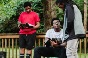 Photo of Williams' sons reading different books. One standing in a red shirt and black shorts and socks, another sitting in a white shirt and black pants, and another looking down at the one who is sitting with lots, a blue sport jacket, and khakis. They all have natural hair and glasses.