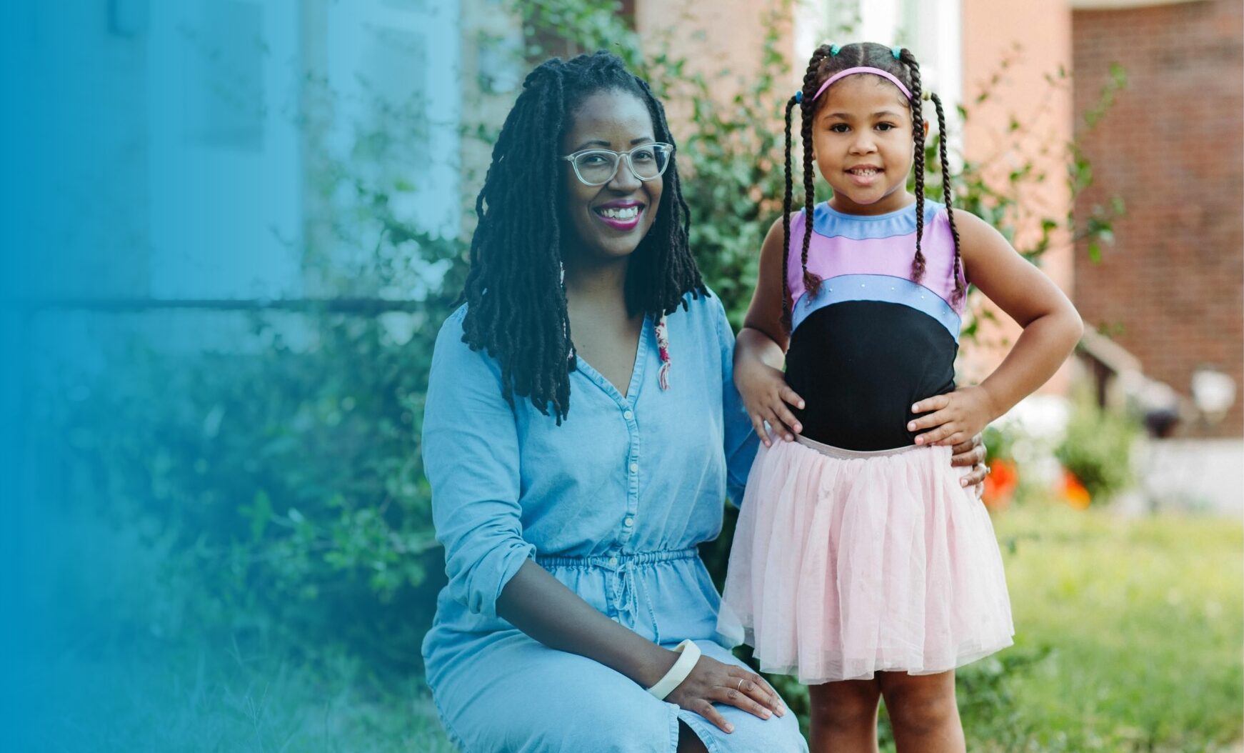 Photo of Amanda Colón-Smith and her daughter. Amanda is a Black woman with glasses, a denim dress, and twists. Her daughter is a lighter skinned Black girl with her hands on her hips, a pink tutu, and a top that has black on the tummy and blue and pink stripes on top. The daughter is wearing several braids and is smiling while Amanda is crouched next to her outdoors in their front yard.