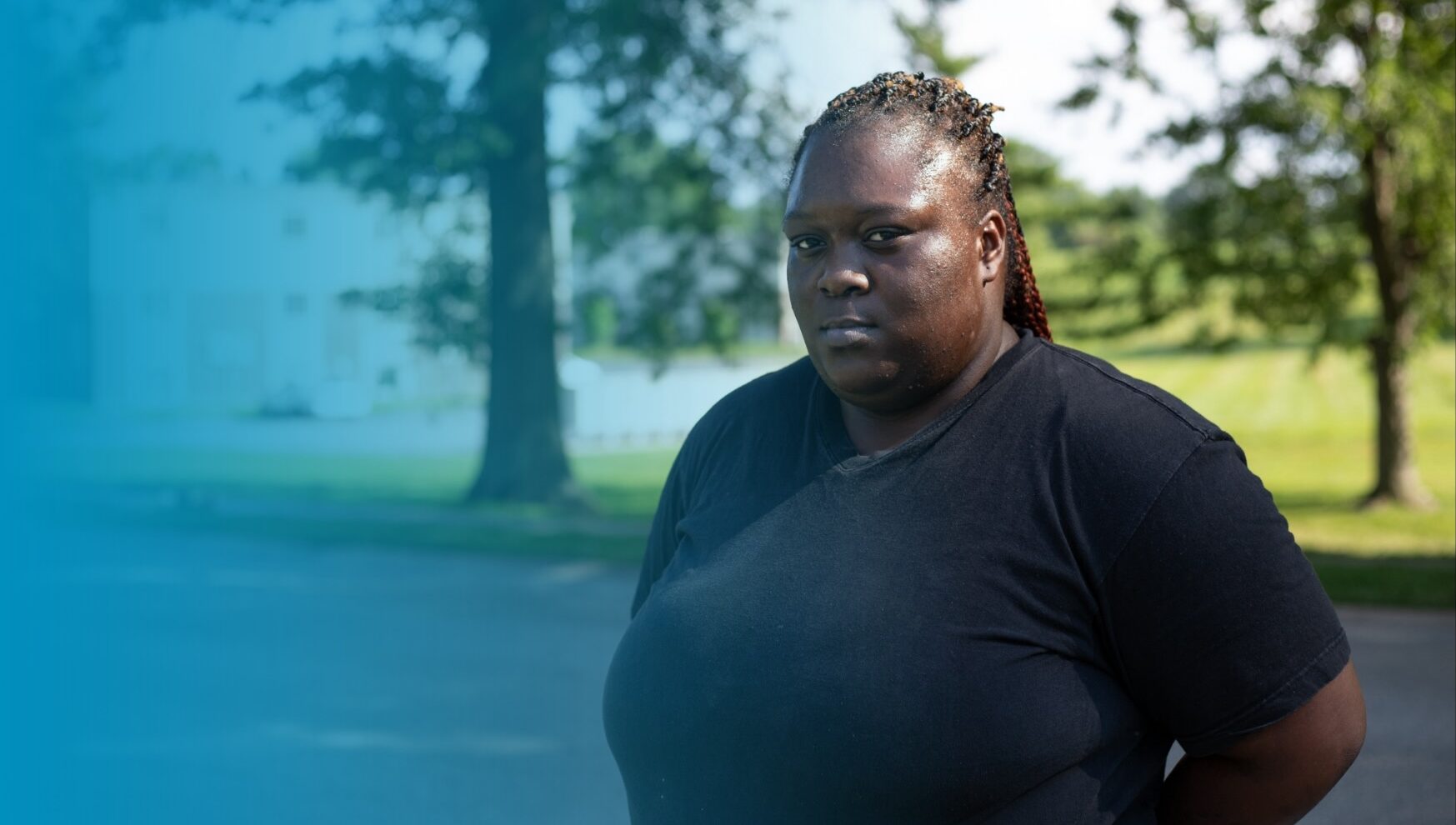 Ebony Williams is a Black woman with braided hair that is pulled back. She has a black t-shirt on and has a closed mouth smile. She seems both friendly and somber. She is standing in the shade of a tree in the Canfield Green neighborhood where Michael Brown Jr. lived.