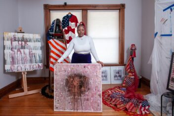 Photo of B.N. BlithedeCarona, a Black mixed-media conceptual artist and resident of Fountain Park. She is holding an art piece entitled Call My Mama Not The Cops. The title is repeated in red ink with a copper and black stain on the front. In the background are other art pieces since as For Sale 100 % Cotton, an American flag hijab with safety pins, and a collage piece. Bee is wearing a long sleeved white shirt, is bald with bleached eyebrows, glasses, and has a serious expression on her face.