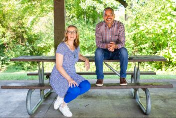 Photo of Dwayne T. James and Toby Weiss sitting together on a picnic bench. In the background is bright sunlight foliage. Toby is a white woman with bright blue pants and white shoes under a blue patterned dress. She has blonde hair that is pinned up and glasses. Dwayne is a Black man with salt and pepper short hair and facial hair. His hands are clasped on his lap as he is leaning forward. He is wearing jeans and brown leather shoes.