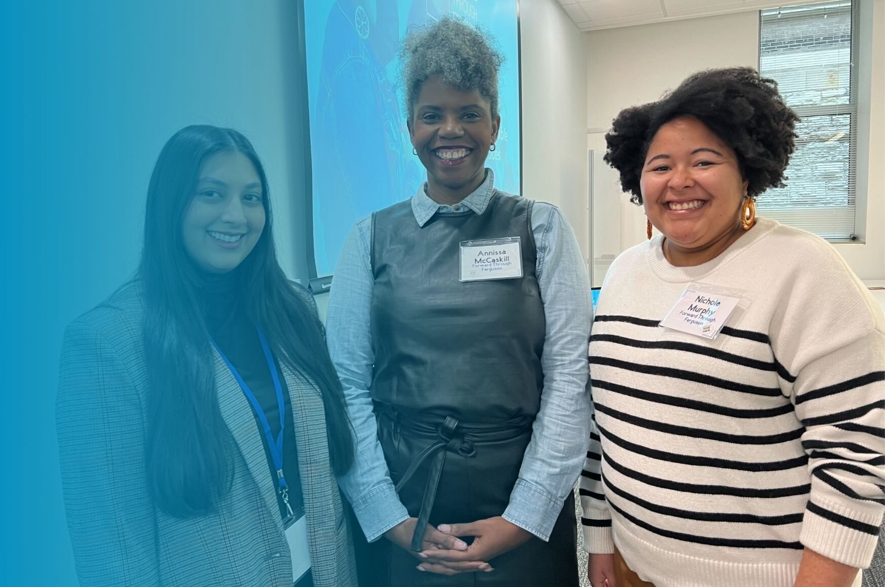 Sara Paracha, Lead Catalyst Resource Development, Annissa McCaskill Executive Director, and Nichole Murphy Director of Community Partnerships are smiling after a presentation about Justice Philanthropy. The presentation is in the background, Sara is a South Asian femme with long black hair and a grey blazer, Annissa is a middle-aged Black woman with grey curly hair piled on her head and a vest over a button up shirt, and Nichole is a light skinned Black woman with a white sweater that has black stripes.