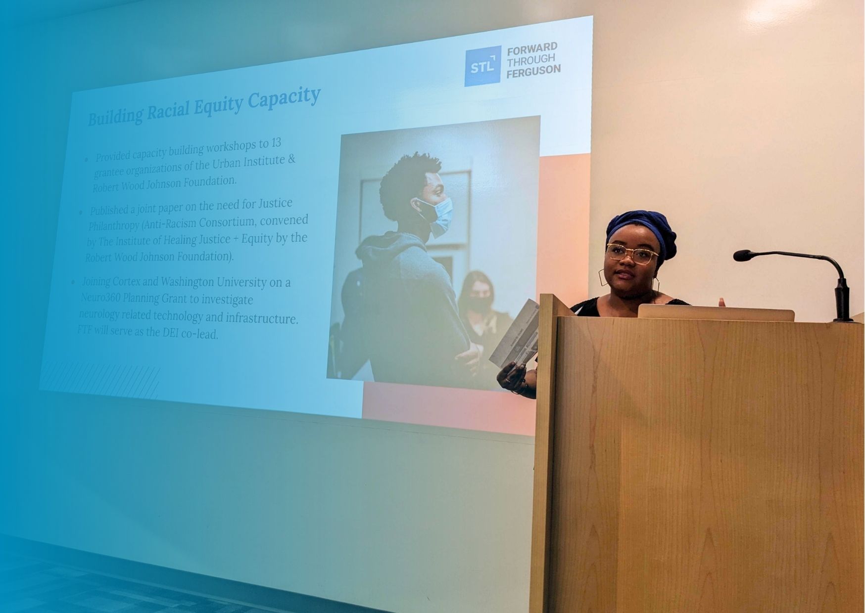 Faybra Jabulani, a Black woman wearing a scarf on her head, gold rimmed glasses, and holding a piece of paper, is standing behind a podium and presenting about building Racial Equity Capacity.