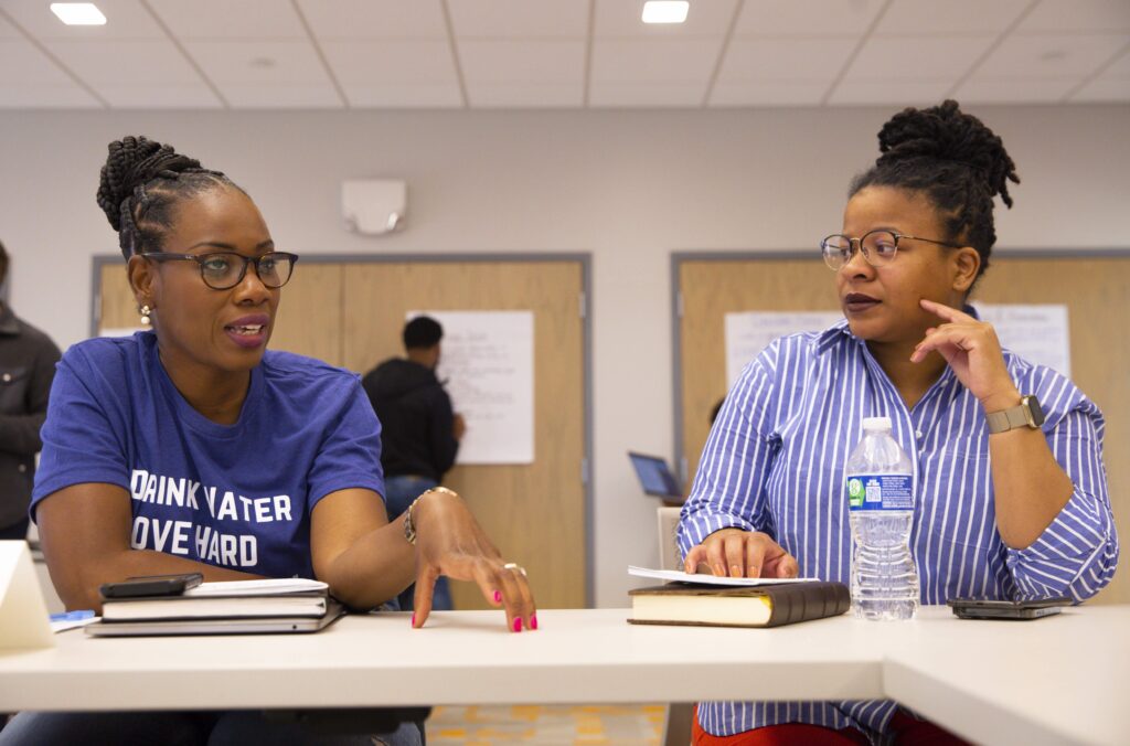 Photo of two CGB members speaking to each other at a table with notes. Both are Black women with natural hair and glasses. One is making a point while gesturing on the table, and another is looking on thoughtfully.