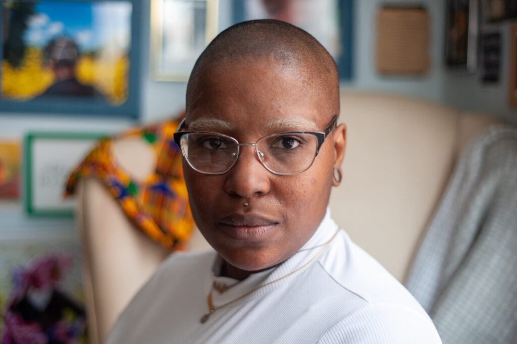 Close-up photo of B.N. Blithe de Carona's face. Bee is a Black artist who is bald, has blonde eyebrows, wears glasses, and is wearing a white long sleeved shirt with golden necklaces. In the background are various photos and pieces of art that are blurry. Bee has ear piercings and a piercing above her lip and looks seriously at the camera.