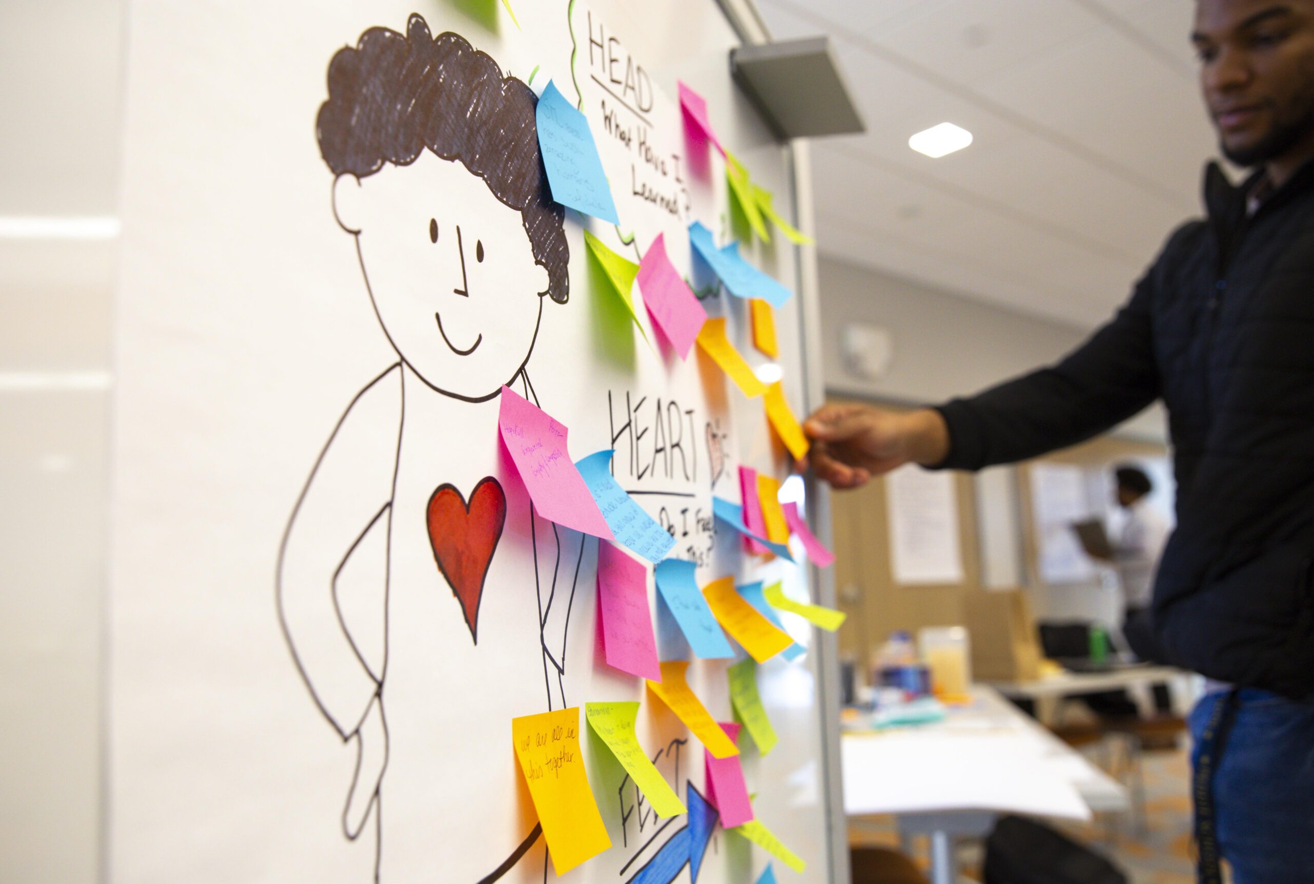 Russell Beckham, former Community Partnerships Catalyst, is placing a post-it on a piece of paper on the wall that has an illustration of a person with a red heart on them. The person has curly hair and is surrounded by colorful post-it notes with ideas.