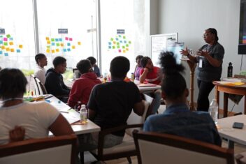 Antionette Caroll, a Black woman in braids, glasses, a grey t-shirt and jeans is speaking to young Black creatives. She is the Executive Director of Creative Reaction Lab. The young people are sitting in desks with colorful post-its on the walls.