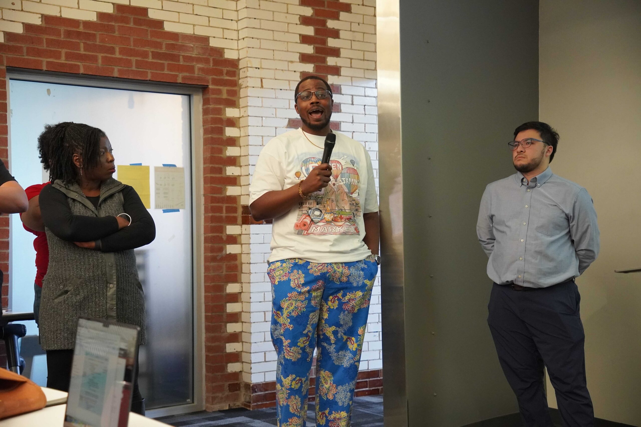 Marcel, a Forward Through Ferguson board member, is a tall nonbinary Black person in a graphic tee, glasses, short hair and short beard, wearing floral blue pants. Riisa and Ishak are two other board members who are listening intently to Marcel speak.