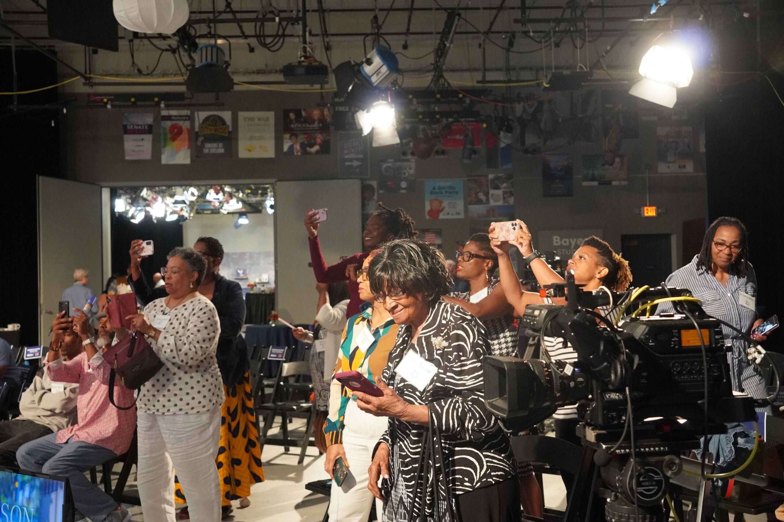 A group of Black people are taking photos on their cell phones in a Nine PBS set for the 10 year anniversary of the Ferguson uprising Listen St. Louis podcast by Carol Daniels. There are bright lights and everyone is smiling. There are people old and young and large TV cameras.