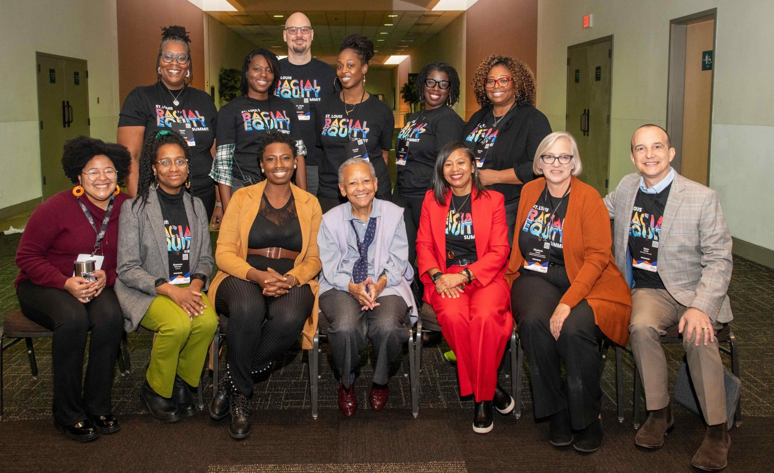 A group of volunteers for Racial Equity Summit 2023 are surrounding esteemed poet and elder, Nikki Giovanni. They are a group of Black and white volunteers who are smiling, many of whom have Racial Equity Summit t-shirts.