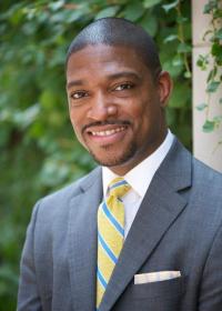 Rev. Dr. Starsky Wilson is a smiling Black man with short hair, mustache and goatee with a gray suit, white shirt, and yellow tie with teal stripes. There is blurred foliage in the background. 