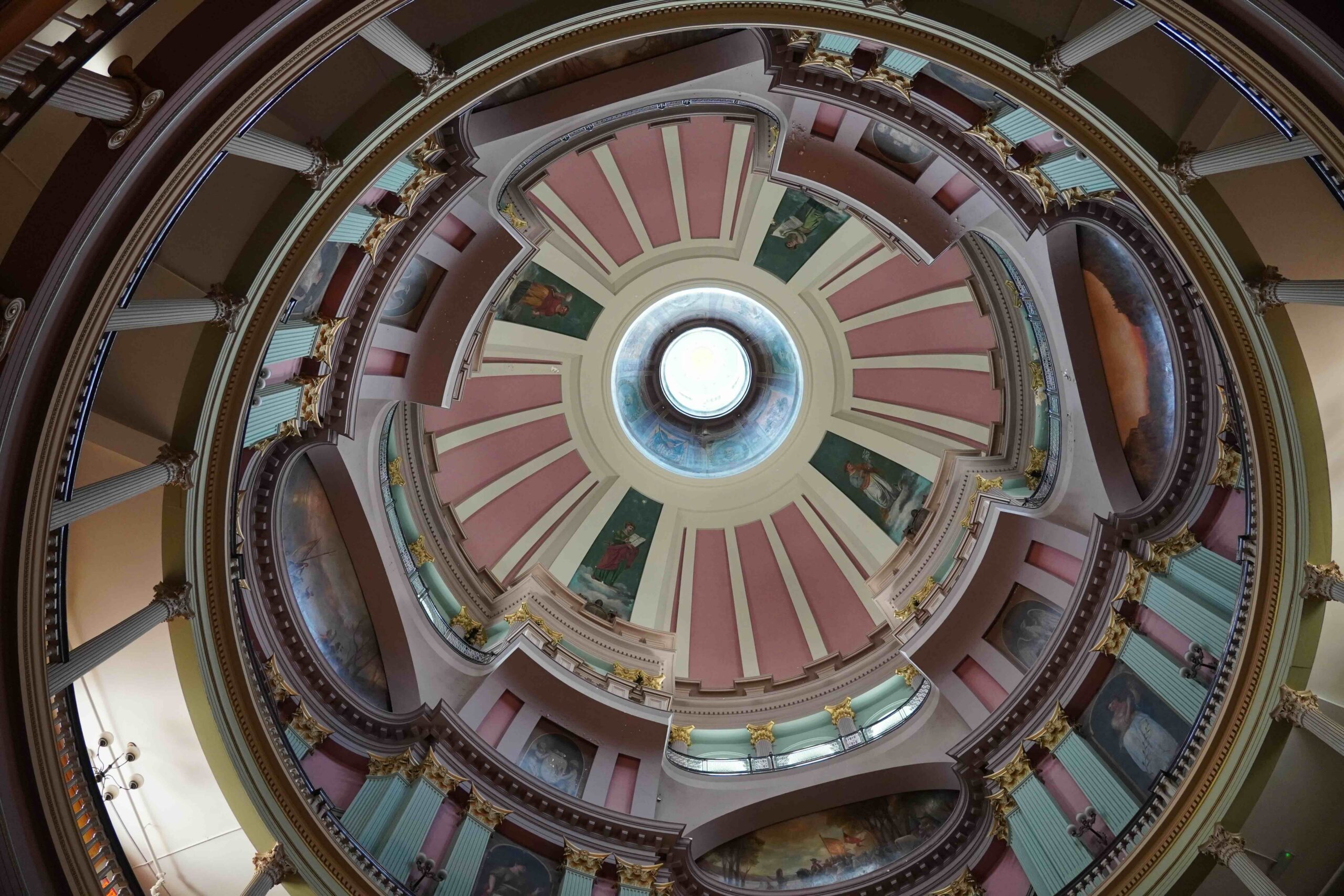 The inside view of the ceiling Old Courthouse. It is ornate with rings and levels of paintings interspersed with coral, yellow, seafood green panels and details. You can see the skylight above.