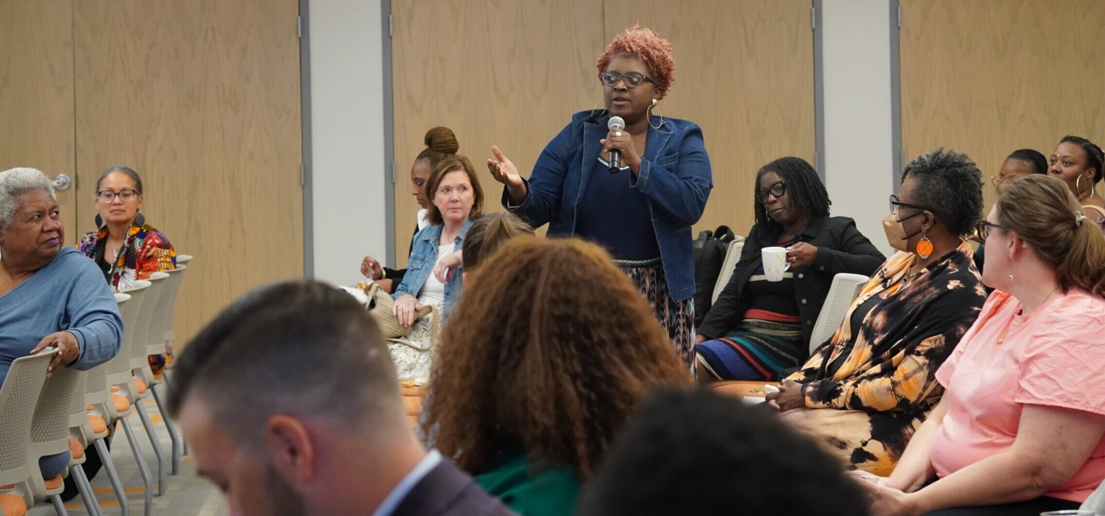 A Black woman with orange short hair and glasses wearing a jean blazer is speaking into a microphone as a diverse group of people pay attention during a Racial Healing + Justice Fund community update.