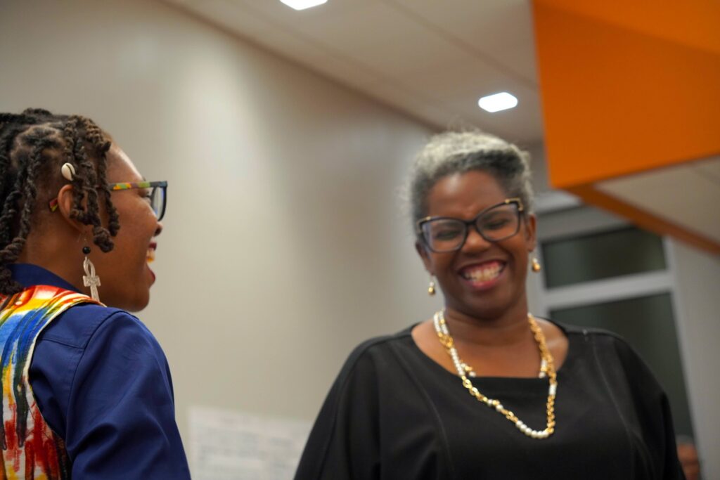 Annissa and Dr. April are laughing together, they are two Black women, one older with grey hair and one younger and middle aged. Dr. April has twists, glasses, and colorful clothing. Annissa has her hair up, with gold jewelry and a black top.