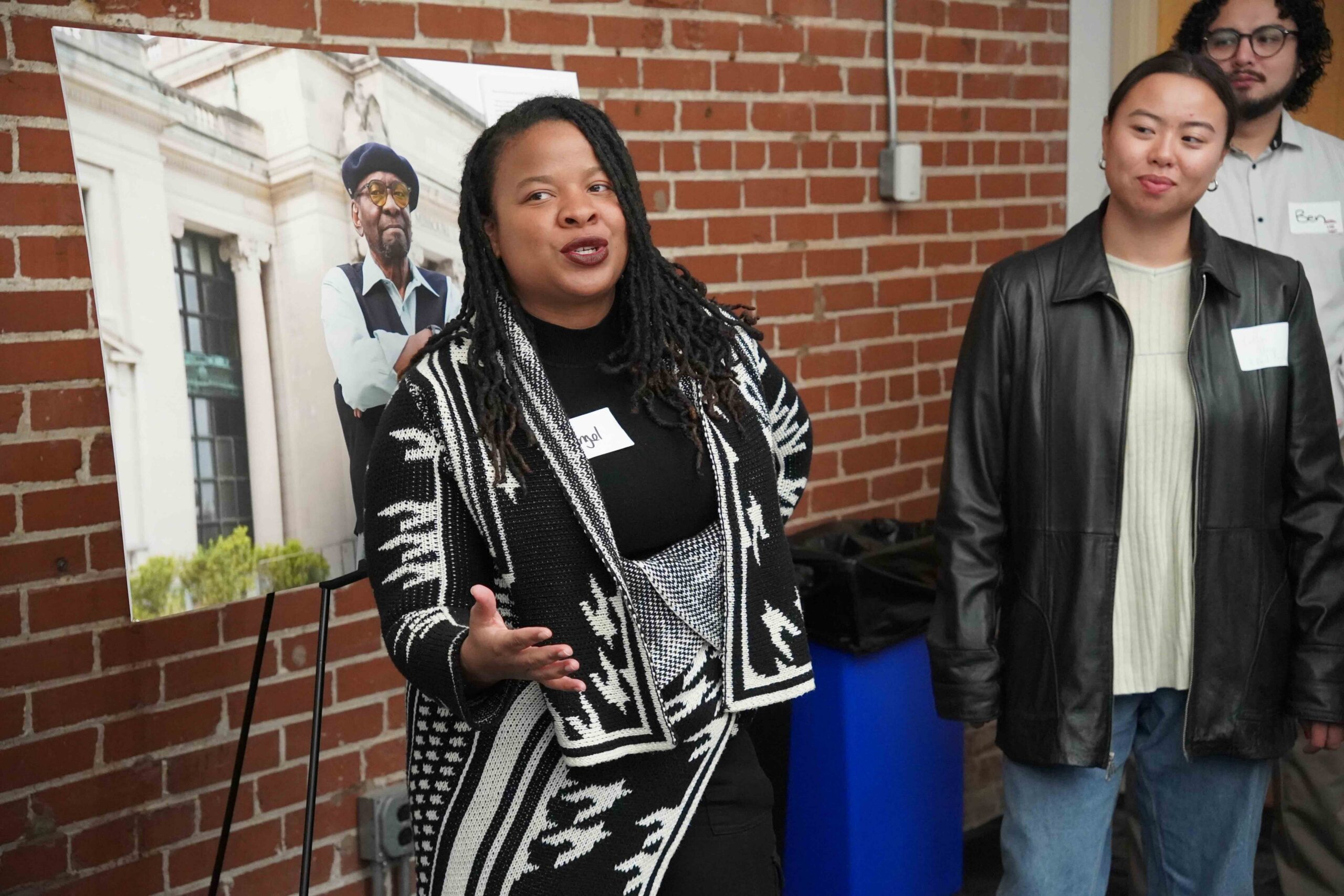 Angel McCain is a Black woman with twists, a black shirt, and a black and white patterned cardigan who is speaking emphatically. Looking on are other people who attended a community update and in the background is a photo of Percy Green, a civil rights icon in St. Louis who is standing in front of of the history museum.