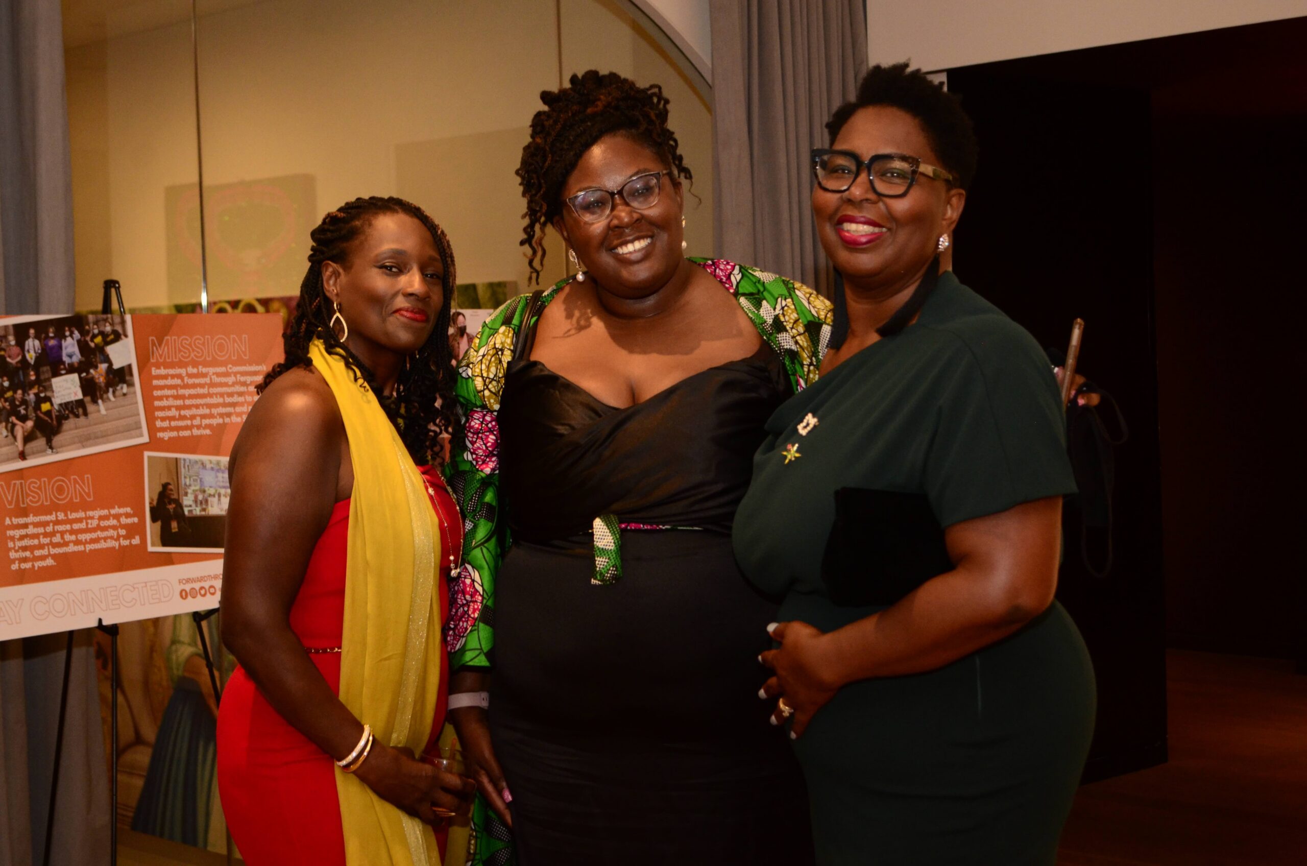 Three Black women, one with twists, a red dress and yellow scarf, another with hair up and black dress with colorful blazer, and another with short hair and emerald dress have their arms around each other. Two of the women are the daughter and mother, Nicole and Adeola Adewale. They are smiling and glowing at the unflinching progress fundraiser for Forward Through Ferguson.