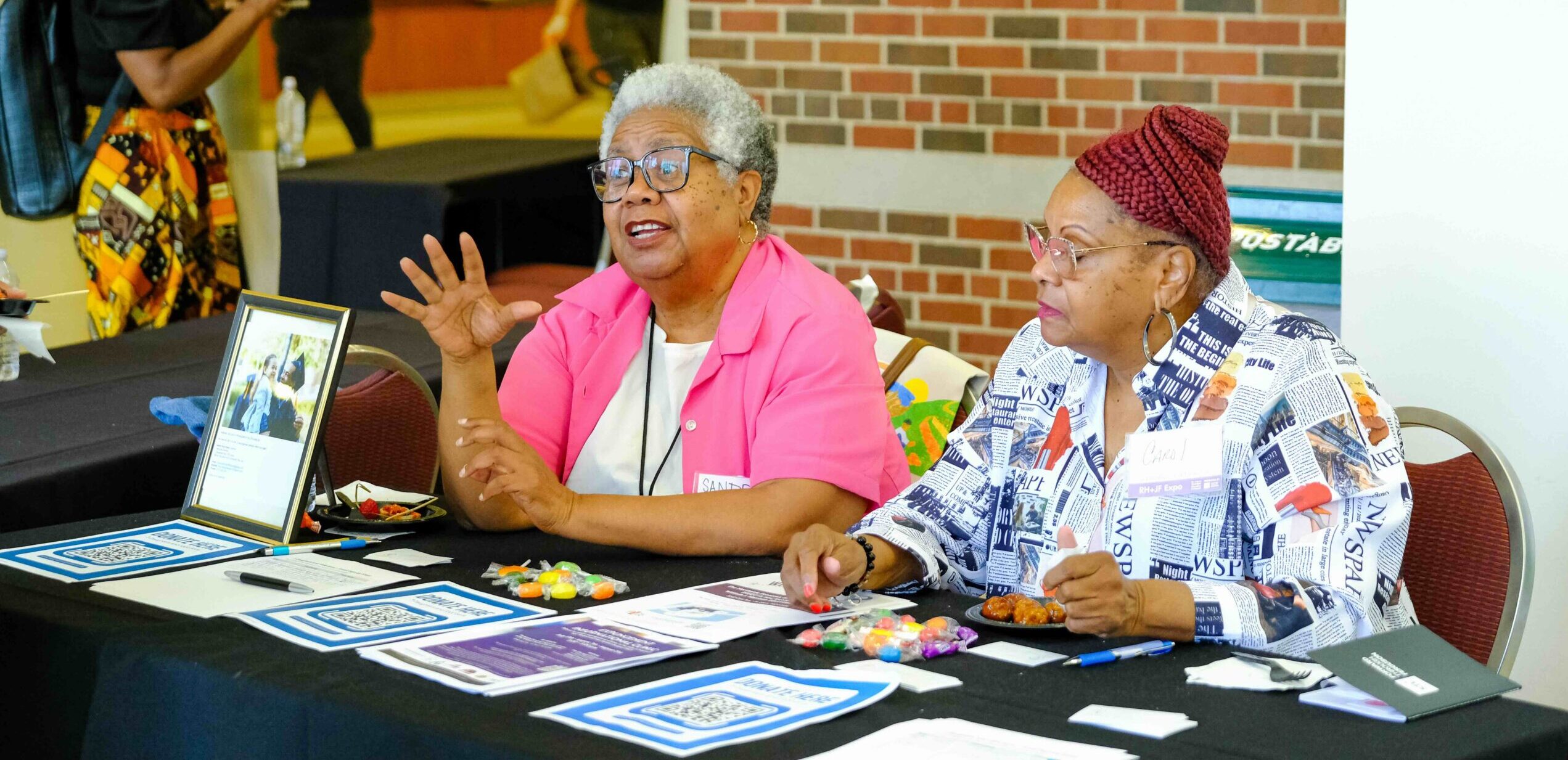 Sandra Keely is an older Black woman with short white and grey hair and glasses, wearing a hot pink button up shirt over a white t-shirt is gesturing as she speaks to someone at her table at Racial Equity Summit. She is sat next to someone else who volunteers with the organization, an older Black woman with red braided hair, glasses, and a colorful patterned button shirt.
