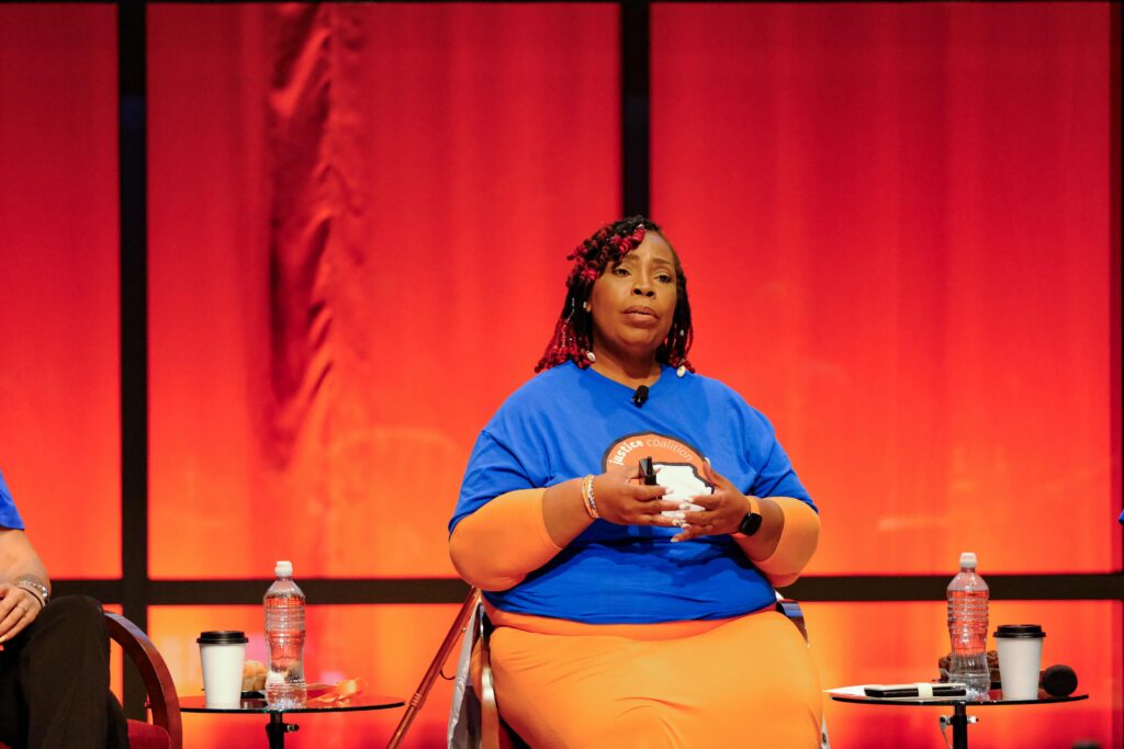ML Smith from Missouri Justice Coalition talking about her work to abolish the death penalty and advocate for people on death row. She is a Black woman with black and red locks in twists. She is speaking passionately in a blue t-shirt with a logo with the Missouri state and an orange skirt. Her background is a red curtain.