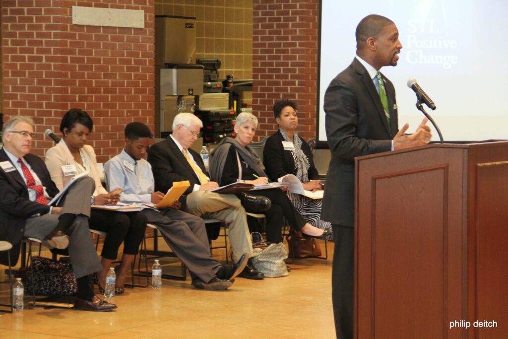 Rev. Dr. Starsky Wilson speaks from a podium as Black and white members of the Ferguson Commission sit behind him.