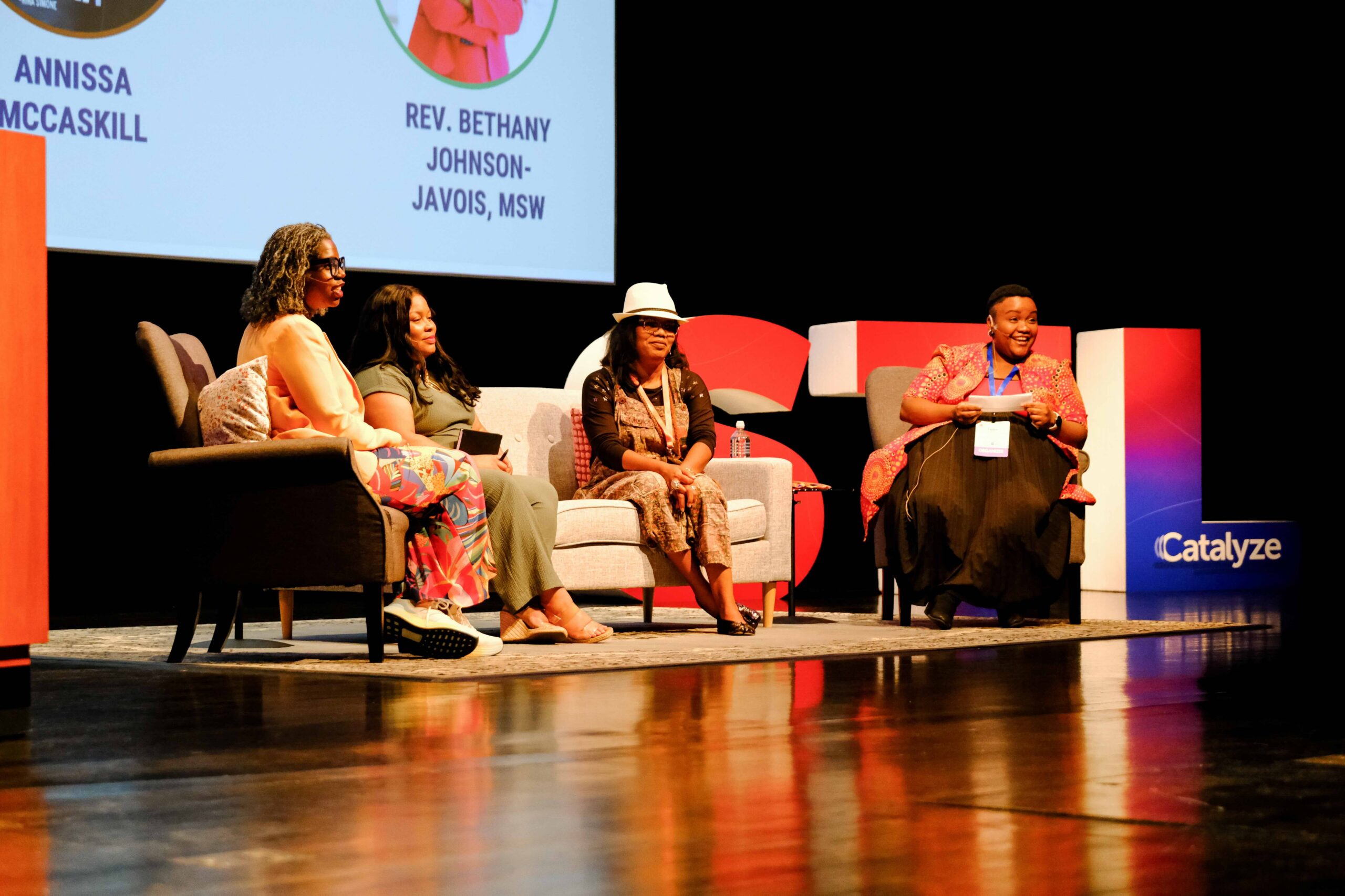 Annissa McCaskill, Constance Harper, Bethany Johnson-Javois, and Faybra Jabulani speak together on stage at the Racial Equity Summit. They are Black woman with varying styles and ages. They are wearing colorful clothing and are passionate and knowledgeable about what they are saying.