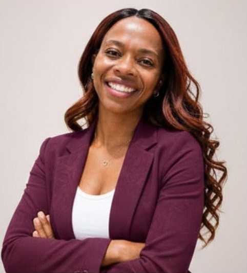 Genesis Glover is a smiling Black woman with lighter skin and long hair with loose curls. She has a beaming smile and is wearing a burgundy blazer over a white tank top with her arms crossed in a confident pose.