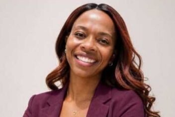 Genesis Glover is a smiling Black woman with lighter skin and long hair with loose curls. She has a beaming smile and is wearing a burgundy blazer over a white tank top with her arms crossed in a confident pose.