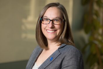 Leslie McClure is a white woman wearing black rectangular glasses and a beaming smile. She has shoulder length brown hair and is wearing a grey blazer with a white tank top.